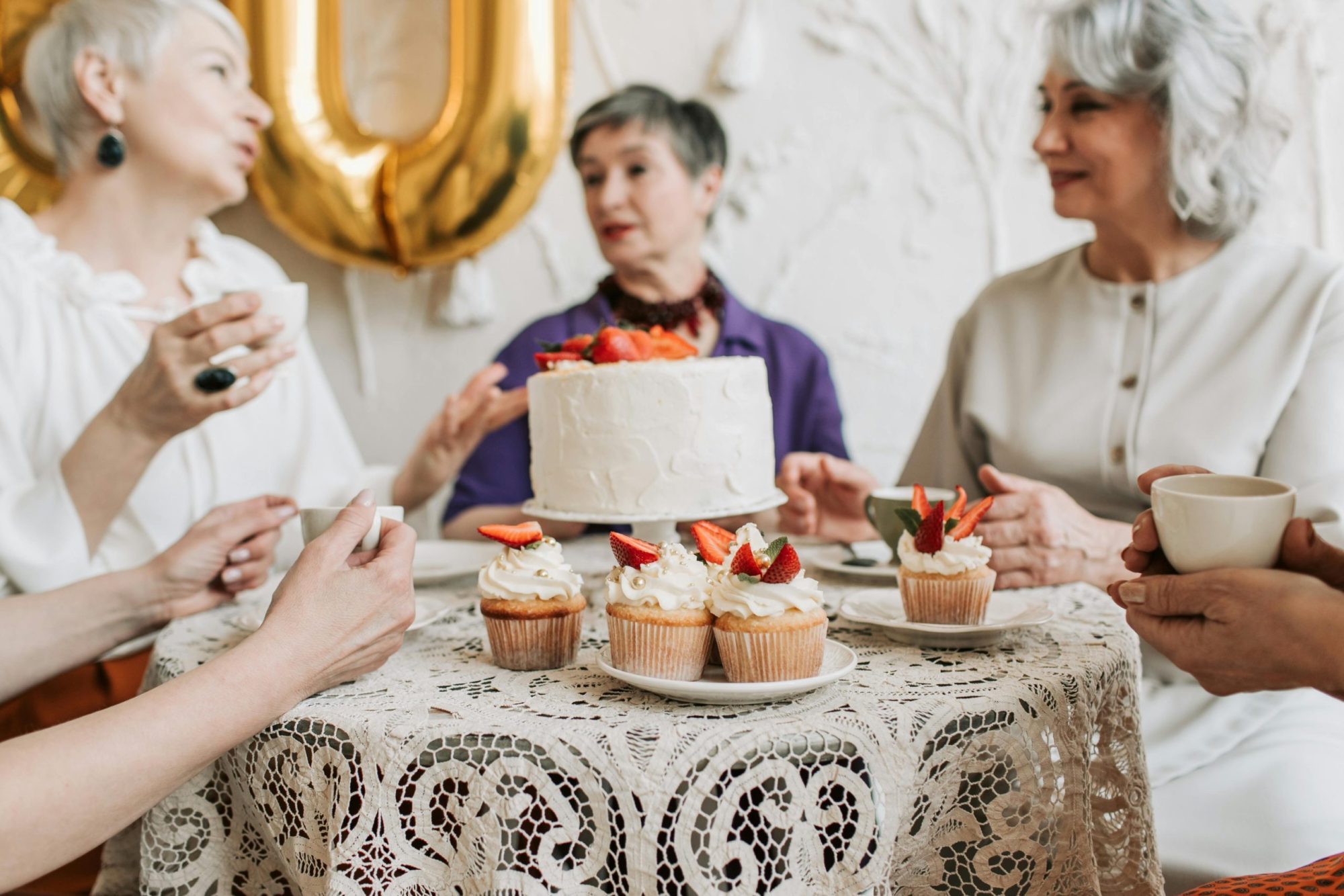 A group of senior women celebrating a birthday with cake and cupcakes indoors.