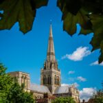 Capture of Chichester Cathedral framed by green leaves against a clear blue sky.
