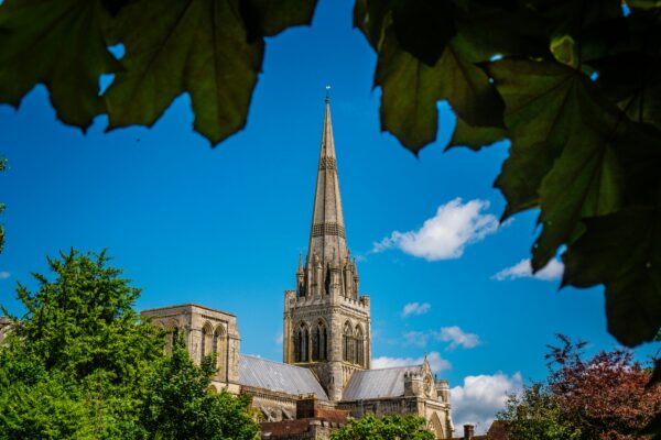 Capture of Chichester Cathedral framed by green leaves against a clear blue sky.