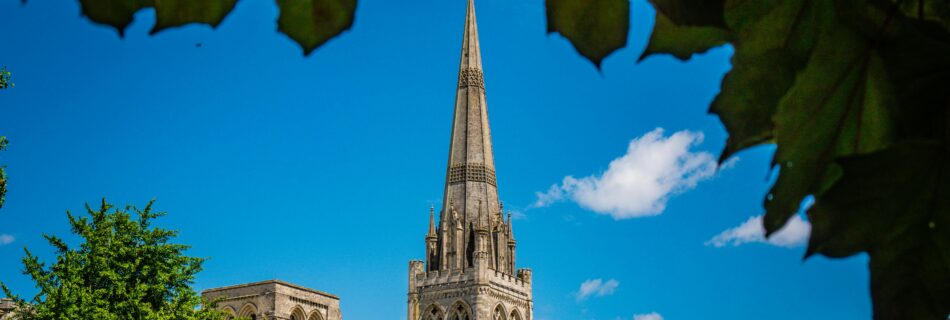 Capture of Chichester Cathedral framed by green leaves against a clear blue sky.